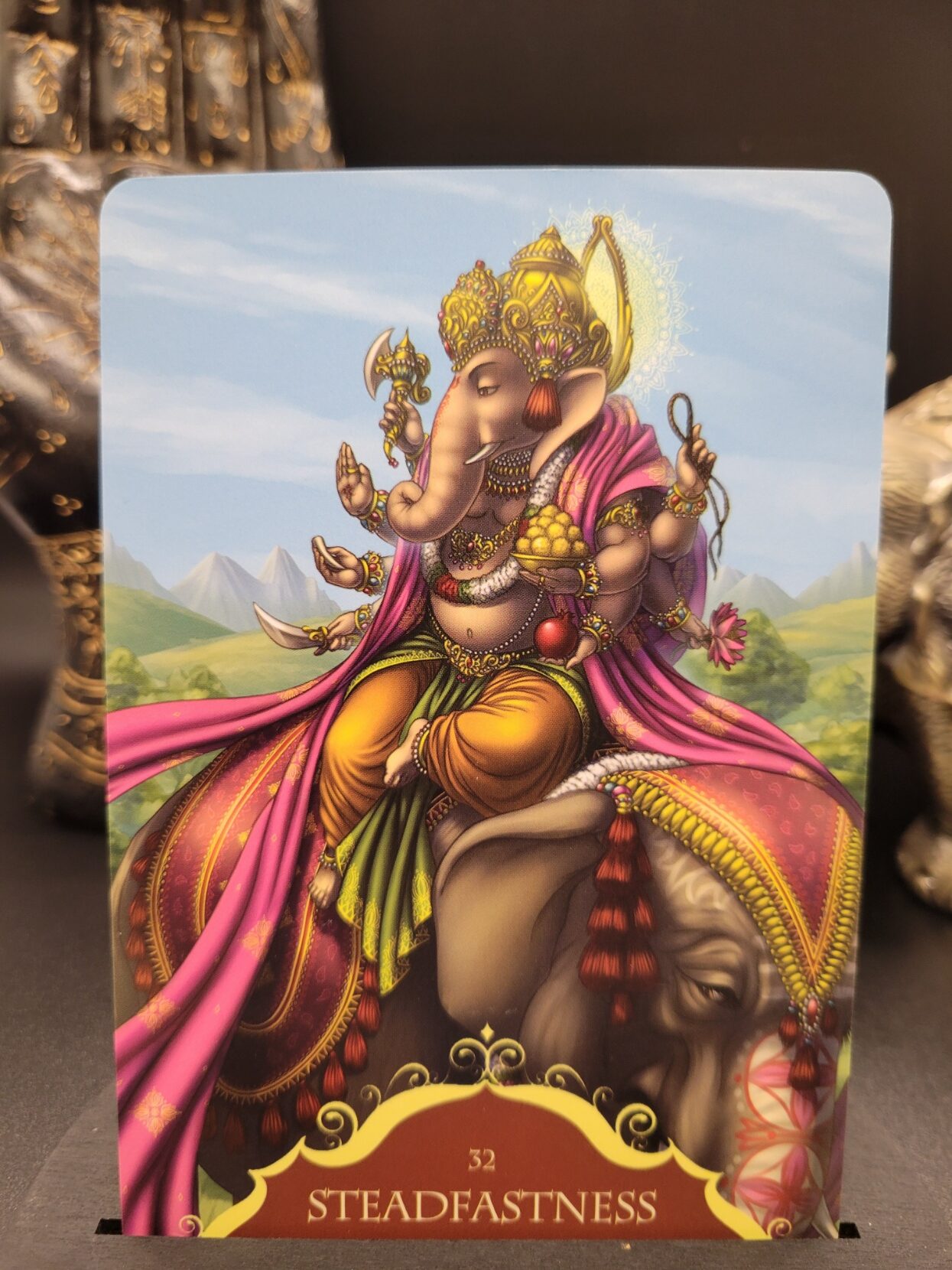 “Steadfastness” oracle card featuring Lord Ganesha seated on a decorated elephant, dressed in ornate gold jewelry and flowing pink robes, with mountains and a blue sky in the background.